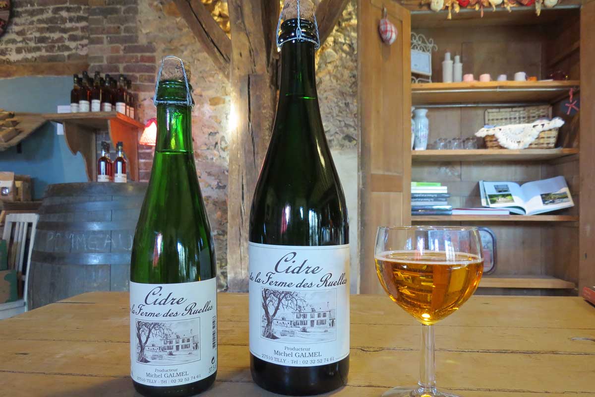 Bouteille de cidre normand de la Ferme des Ruelles, entourée de pommes et de verres remplis de cidre pétillant.