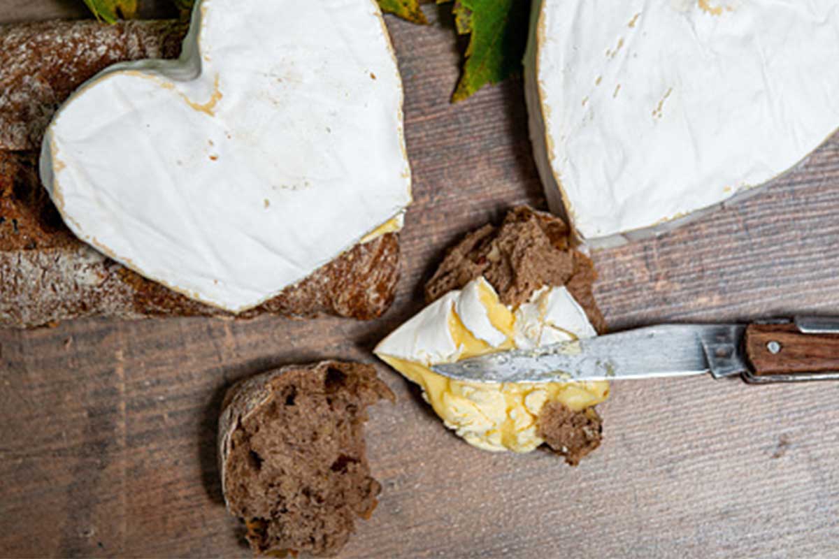 Un fromage Neufchâtel en forme de cœur, posé sur une planche de bois, accompagné de tranches de pain et de raisins.