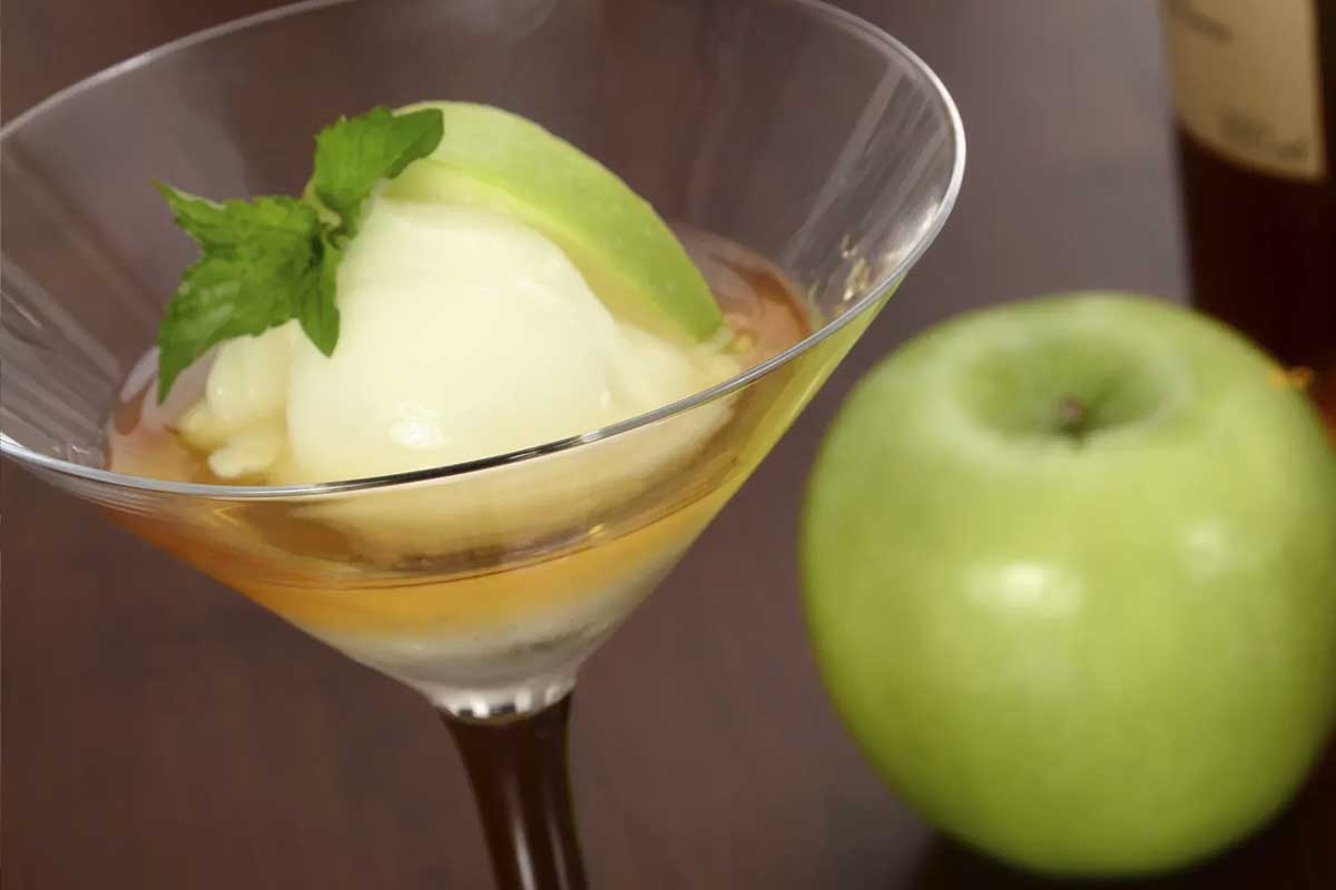 Un verre de Trou Normand, un sorbet arrosé de Calvados, servi dans un verre à pied, avec une bouteille de Calvados et des pommes en arrière-plan.